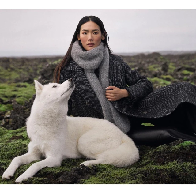 A woman poses outdoors with a white dog. She wears a dark grey herringbone coat and a grey scarf while the dog looks up at her.