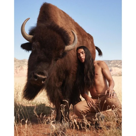 A muscular, Indigenous man poses semi-nude next to a large American bison in a dry, grassy environment beneath a clear sky.