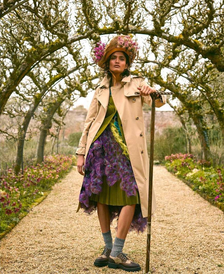An adult female model stands in a garden tunnel wearing a trench coat, patterned dress, hat with flowers, and accessorized with a walking stick.