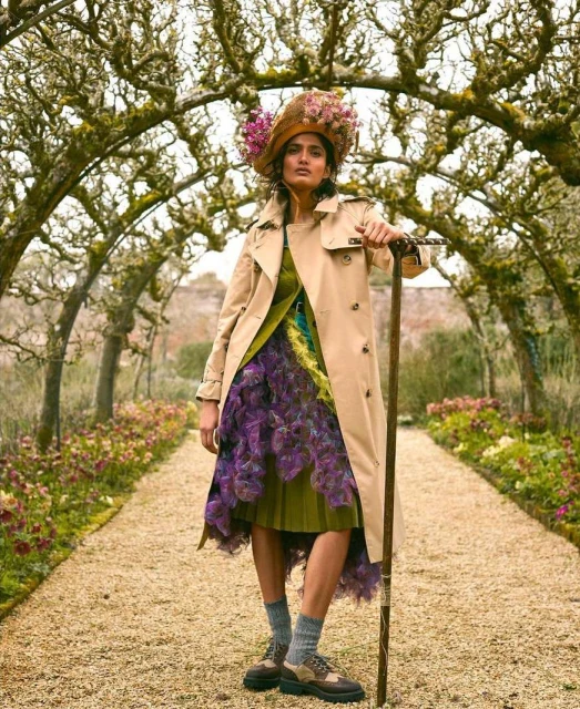 An adult female model stands in a garden tunnel wearing a trench coat, patterned dress, hat with flowers, and accessorized with a walking stick.