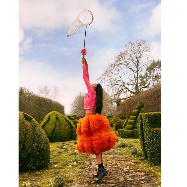 Woman in orange tulle skirt reaches for a butterfly net in a formal garden under a partly cloudy sky, fashion-forward and whimsical.