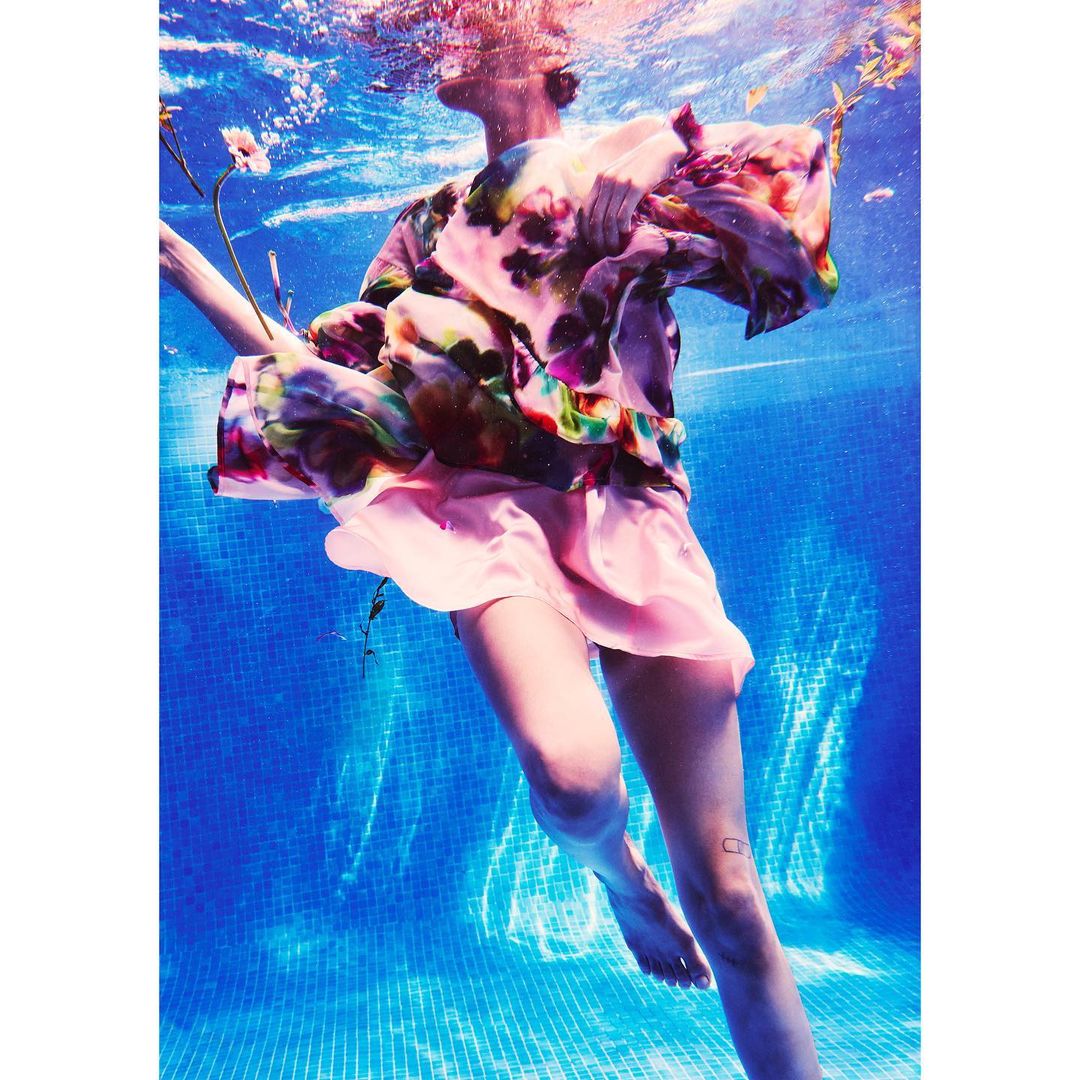 Underwater fashion shot of a woman in a pool wearing a pink dress and floral top, captured in a medium shot with blue water.