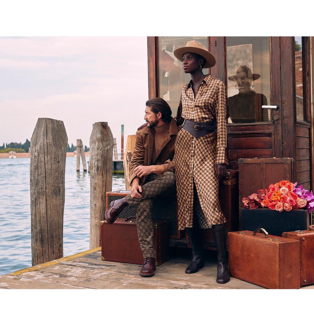 Two fashion models pose on a wooden pier next to vintage luggage and flowers, with a view of the ocean and building in the background.