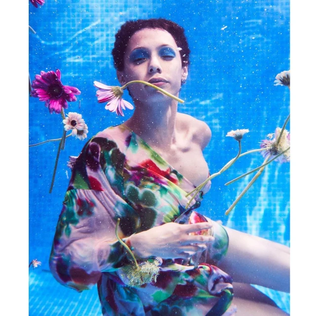 Underwater portrait of a short-haired woman in a patterned dress surrounded by floating daisy flowers in a blue tiled pool.
