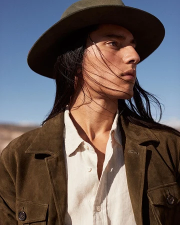 Close-up portrait of a man with long dark hair wearing a fedora hat and olive green jacket against a clear blue sky