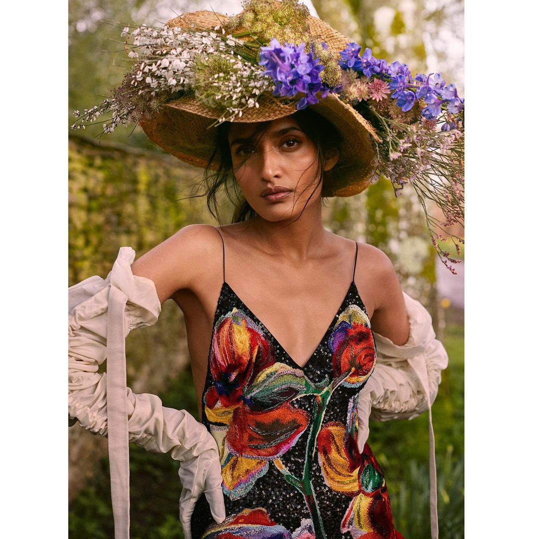Woman in floral dress and straw hat with flowers, posing in a garden setting. Fashion portrait with natural light and blurred background.