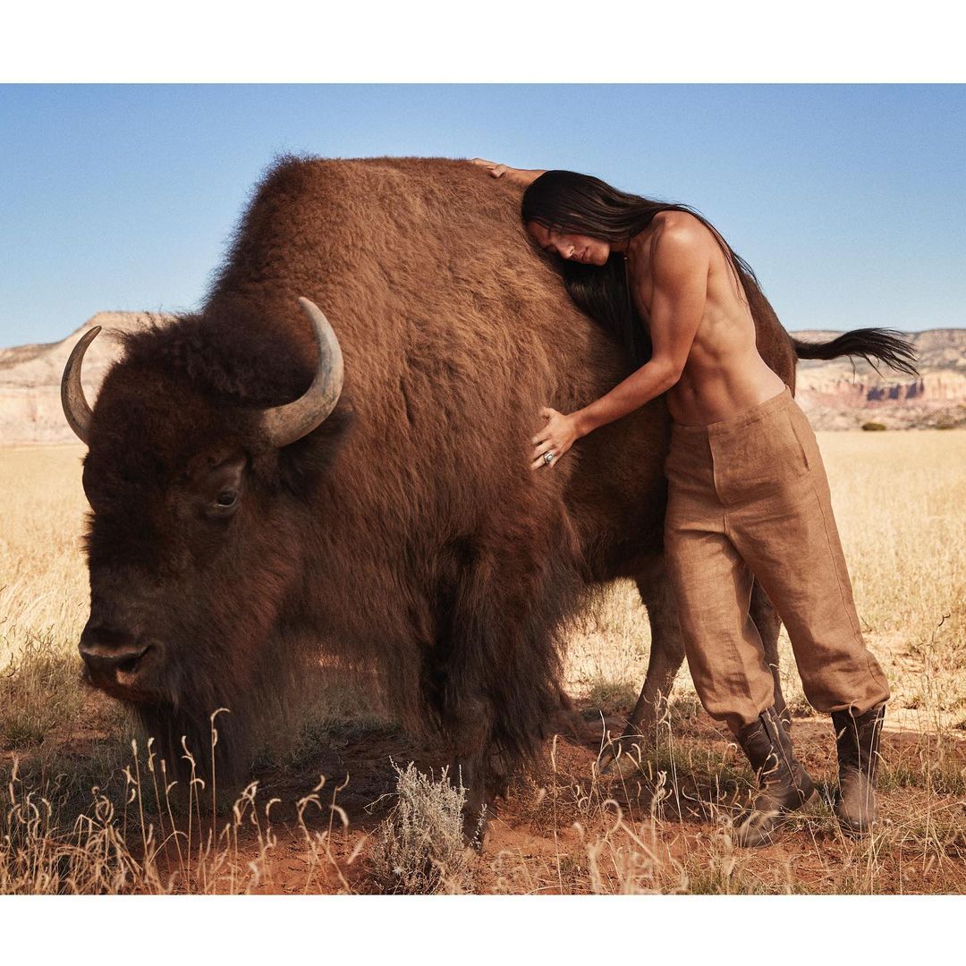 Adult male embraces a bison outdoors in a sunlit, natural setting of dry grass and a clear blue sky, showcasing a unique animal interaction.