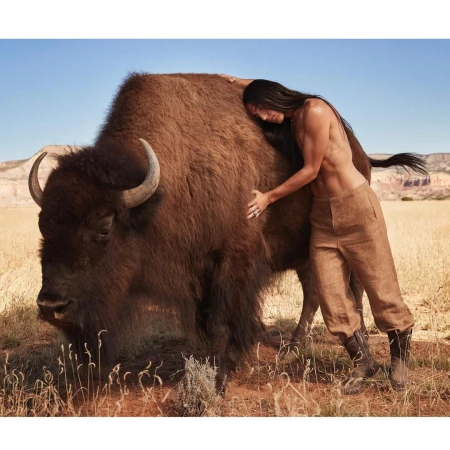 Adult male embraces a bison outdoors in a sunlit, natural setting of dry grass and a clear blue sky, showcasing a unique animal interaction.