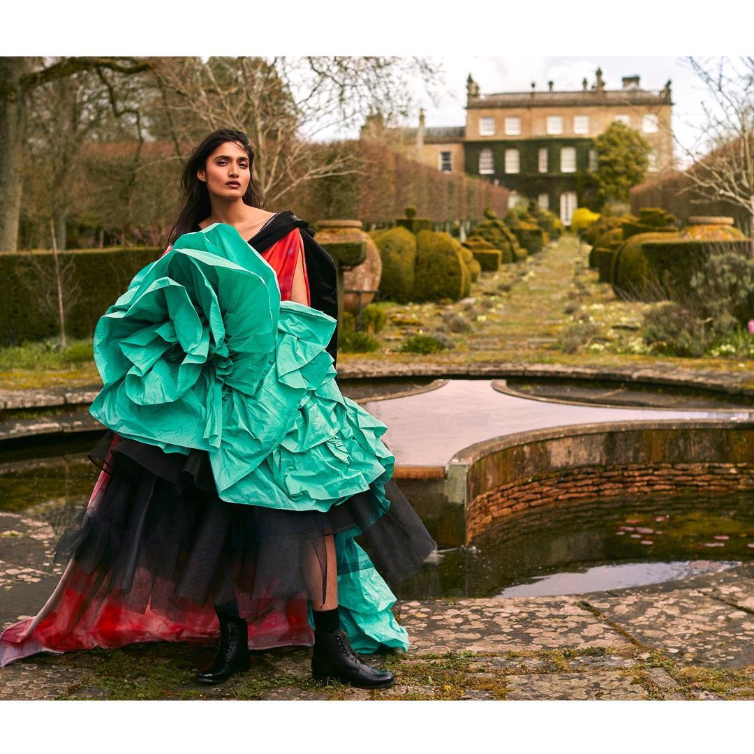Woman in a high-fashion dress with teal ruffles and black boots stands in a garden setting with an elaborate water feature and building.