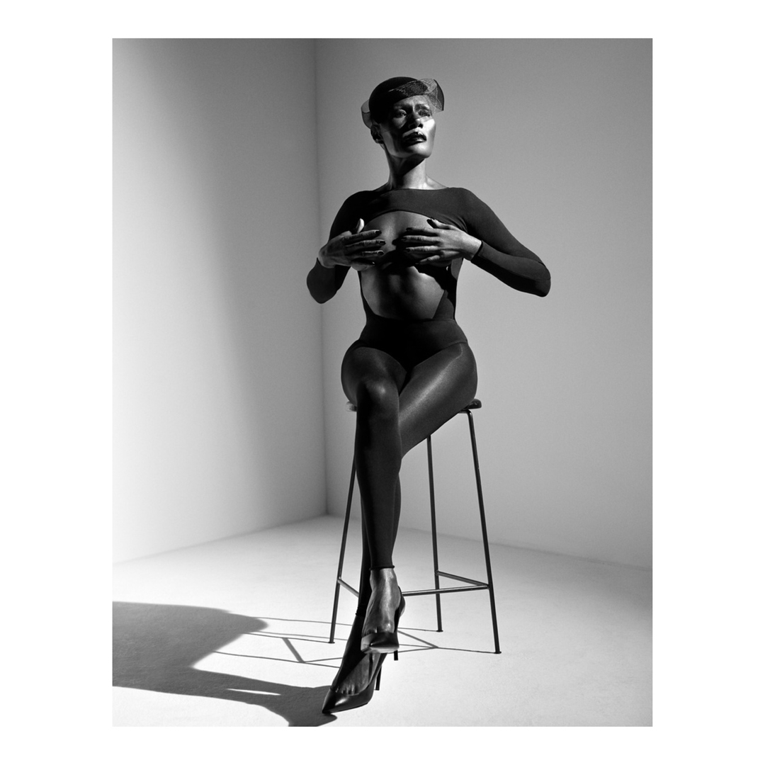 Model wearing dark fitted bodysuit sitting on a tall stool in a studio with directional lighting captured by Richard Phibbs