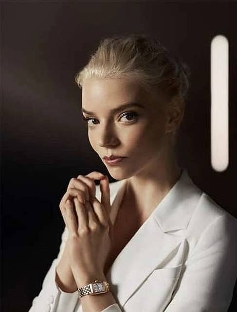 Blonde woman in a white blazer poses with crossed hands, displaying a gold wristwatch against a dark background with a vertical light.