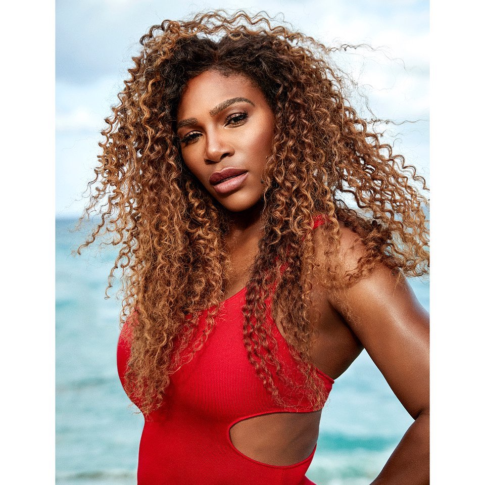 Portrait of a woman with curly brown hair, wearing a red swimsuit, posing outdoors against a blurred background of sky and water.