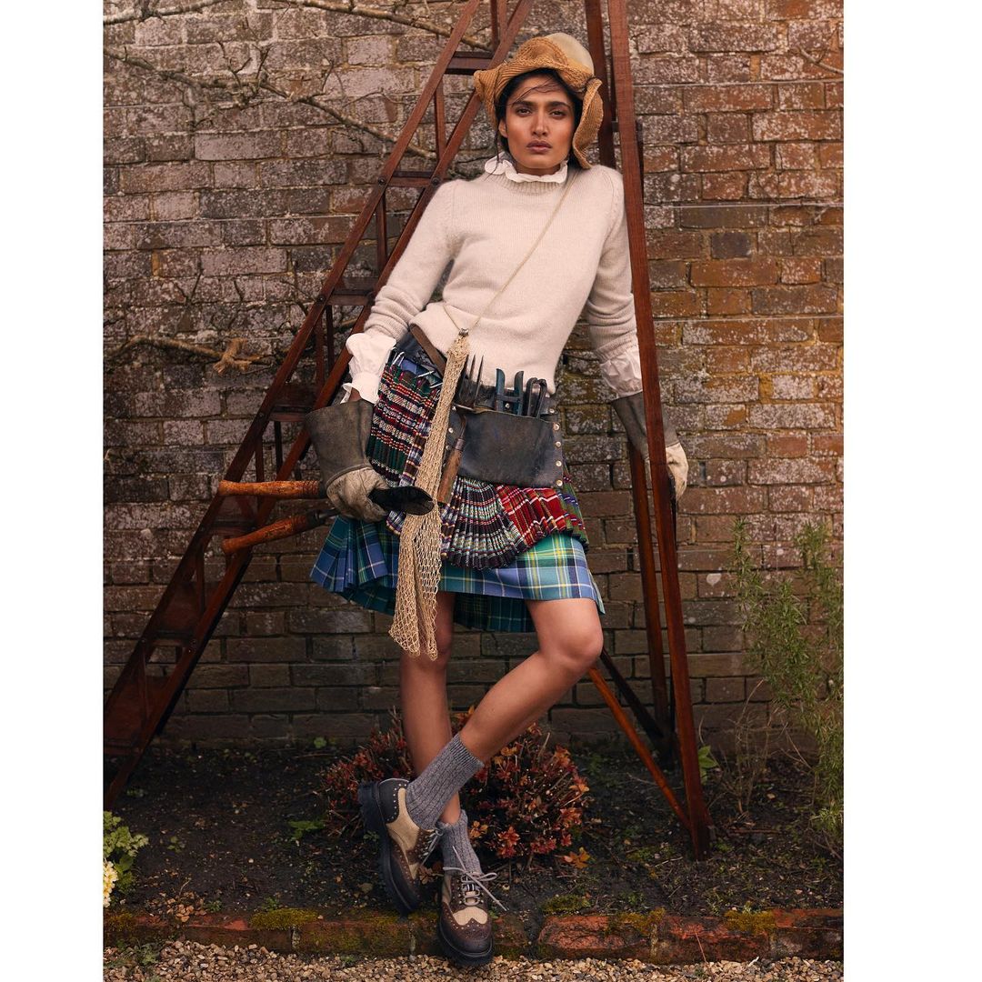 A woman in a kilt and hat stands near a rusty ladder outdoors against a brick wall, creating a vintage fashion editorial image.