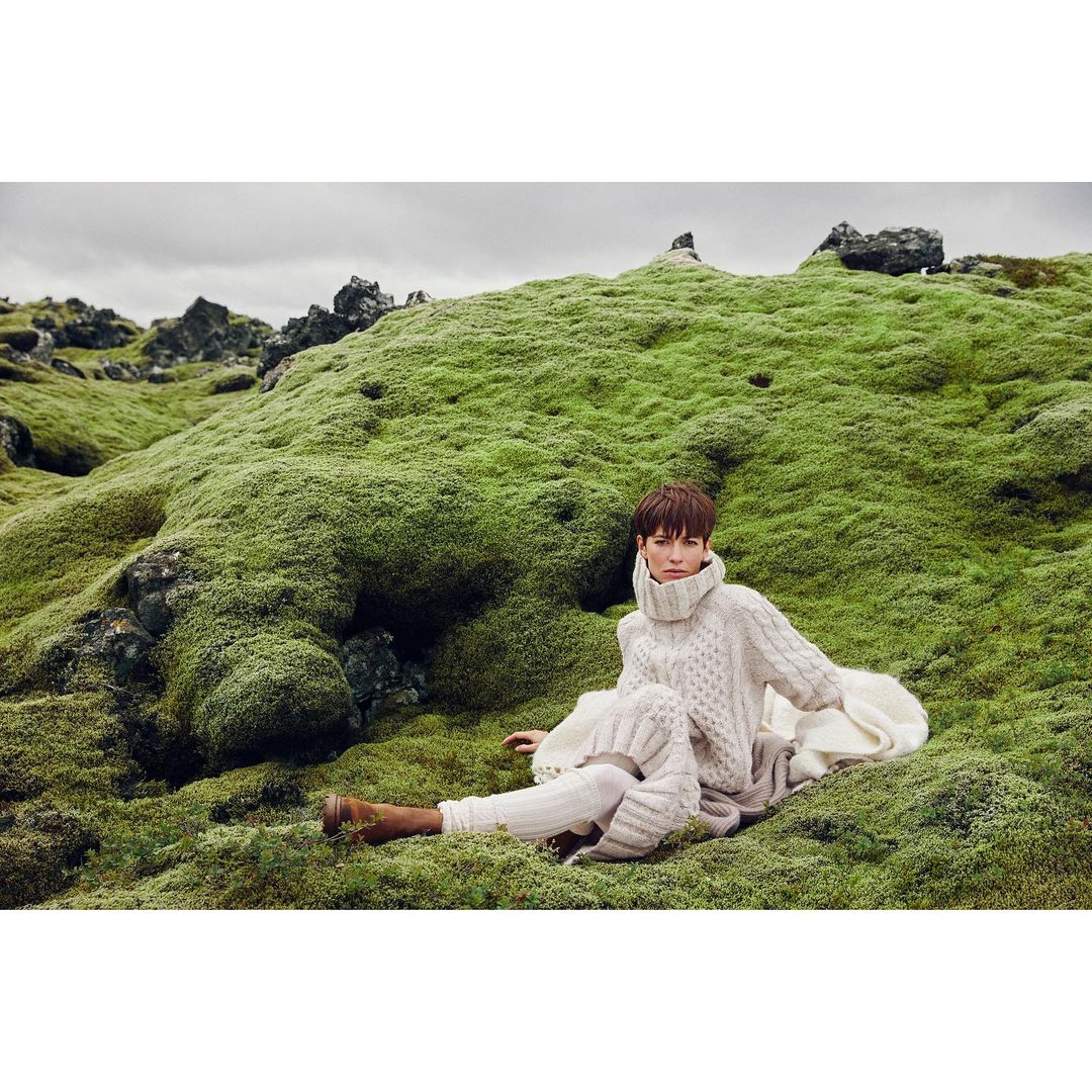 Woman in knitwear sitting on a mossy hillside. She's wearing a chunky turtleneck sweater, wide-leg knit pants and brown suede boots.