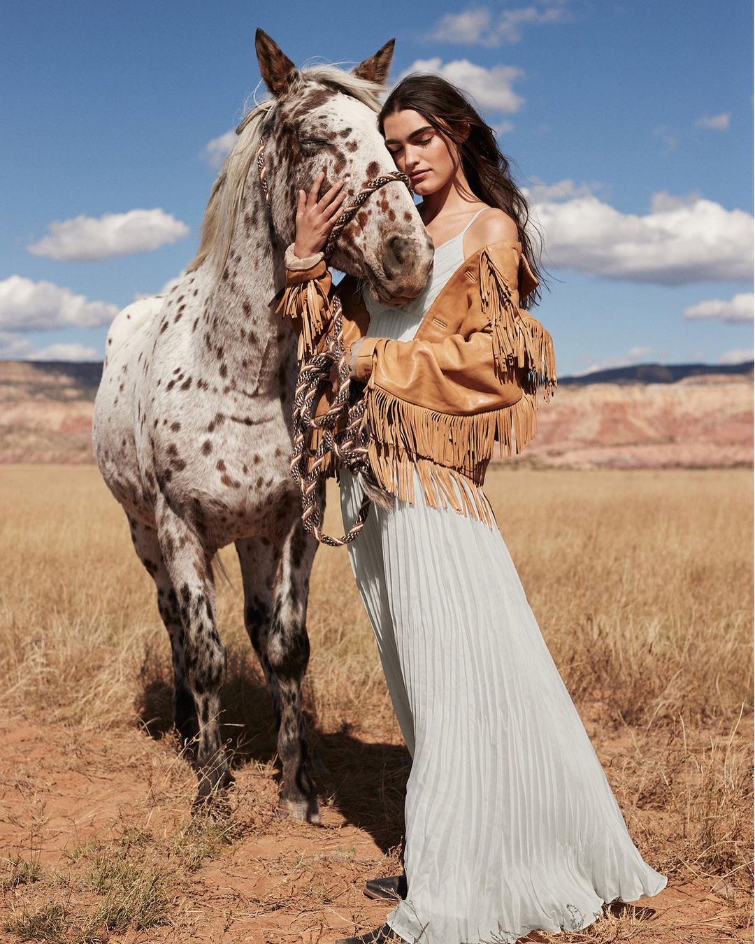 Model in flowing dress poses with an Appaloosa horse in a field with a tan fringed jacket against a blue sky with scattered clouds.
