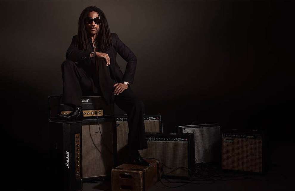 Portrait of a Black adult male wearing sunglasses and a suit, seated on a stack of Marshall amplifiers in a photo studio setting.