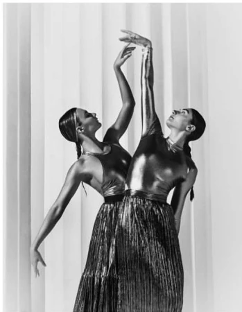 Two ballet dancers in metallic costumes pose together against a white curtain backdrop, showcasing elegance and symmetry in a black and white studio shot.