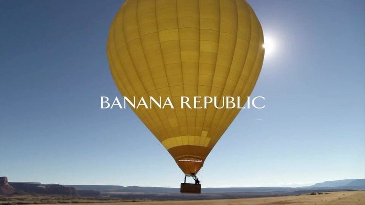 A yellow hot air balloon floats in a clear blue sky over a desert landscape with the Banana Republic logo superimposed in white.