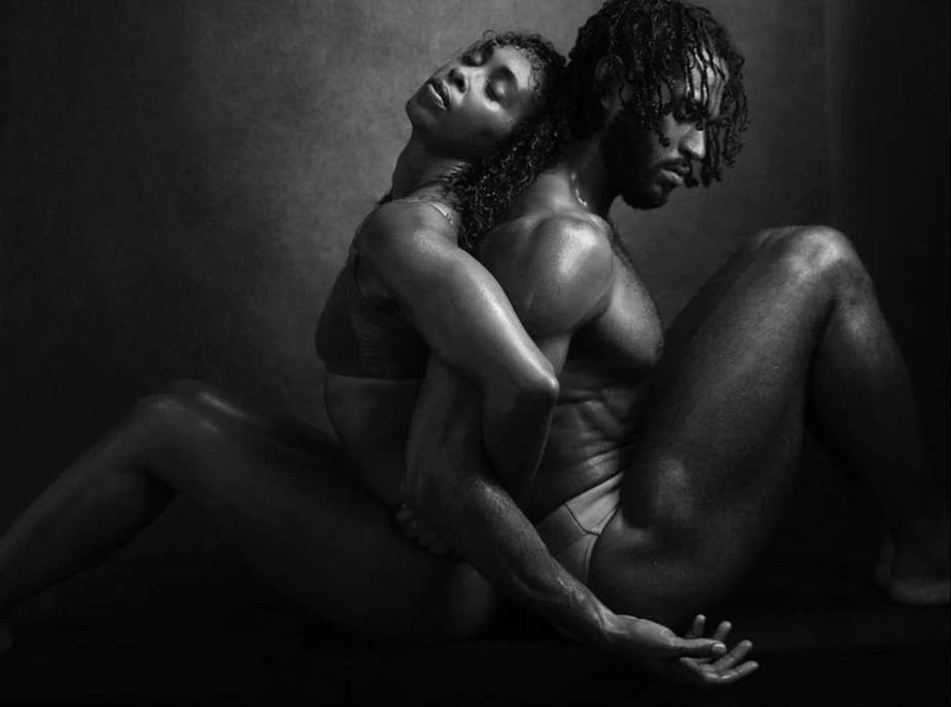 Monochrome portrait of a man and woman embracing, their bodies touching, seated against a dark backdrop in a studio setting.