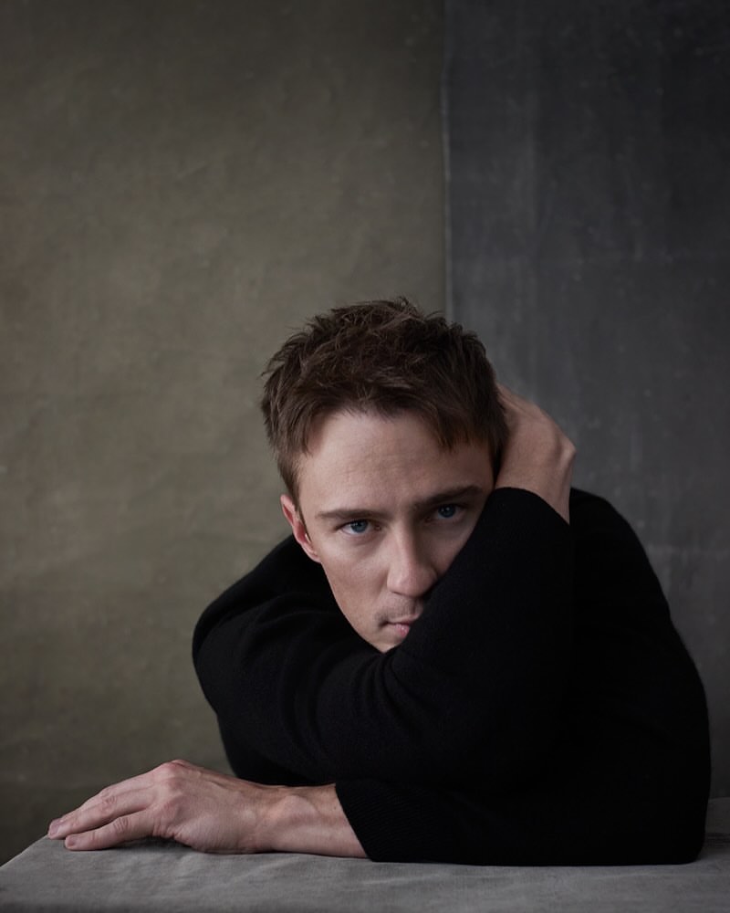 Portrait of a young man with brown hair wearing black top, resting his arms on a grey surface, set against a mottled grey backdrop.