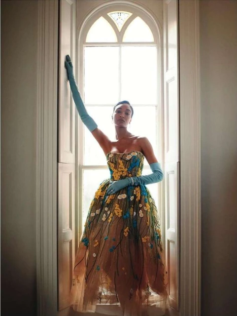 Fashion photo of a Black woman posing in a floral gown by a window, wearing elbow-length gloves; soft, diffused lighting, indoor setting.