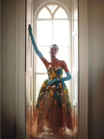 Fashion photo of a Black woman posing in a floral gown by a window, wearing elbow-length gloves; soft, diffused lighting, indoor setting.