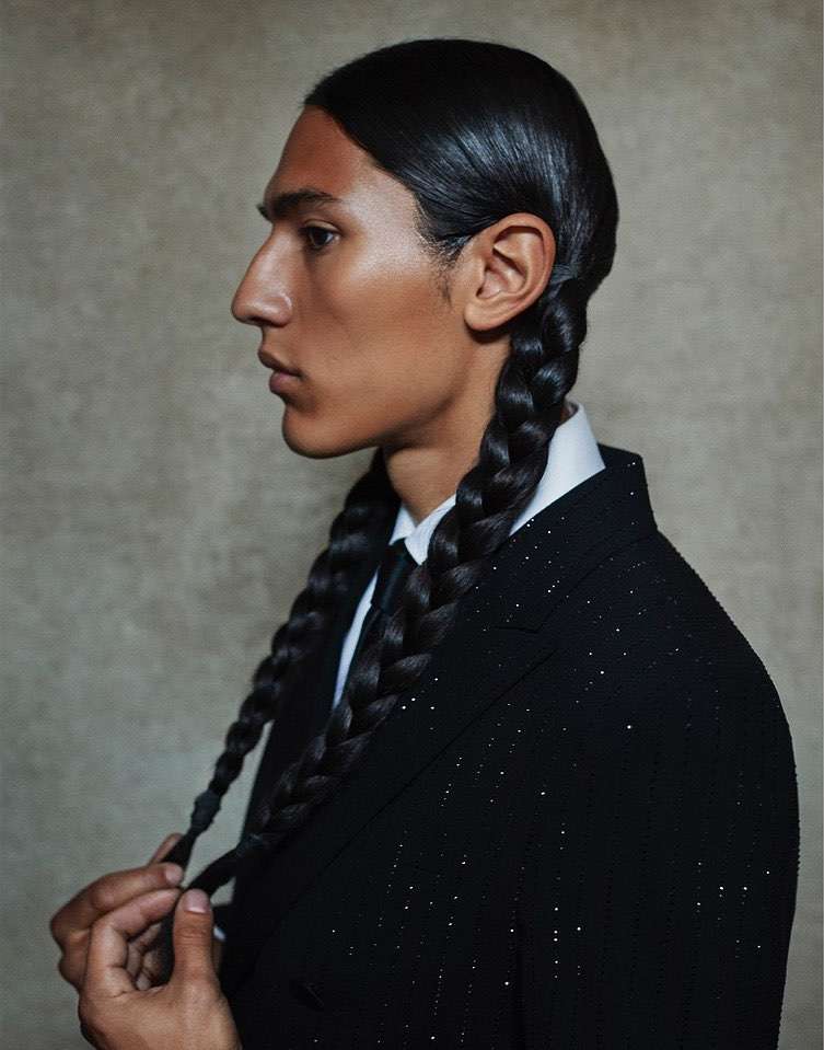 Profile portrait of a dark-skinned male model with long black braided hair wearing a black suit and tie against a neutral backdrop.