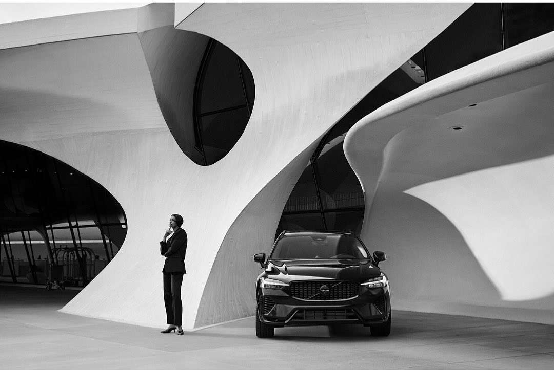 Black and white shot of a black Volvo parked in front of a modern architectural building with an adult female standing to the side.