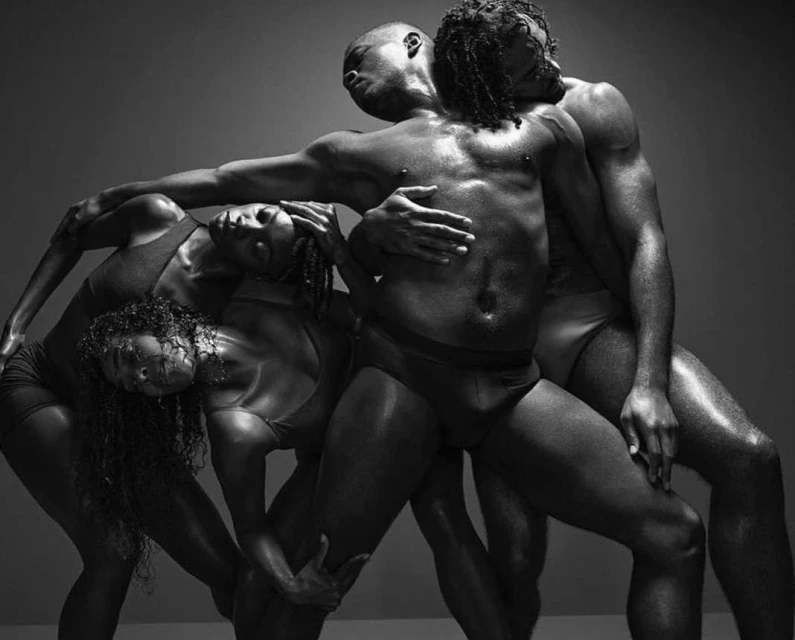 Striking black and white studio image of four athletic African descent dancers intertwining, their bodies glossy with sweat under bright lighting.