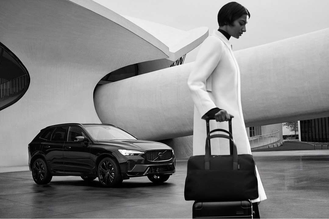 A modern black Volvo SUV and woman with a rolling bag stand before an architectural building in a monochrome outdoor shot.