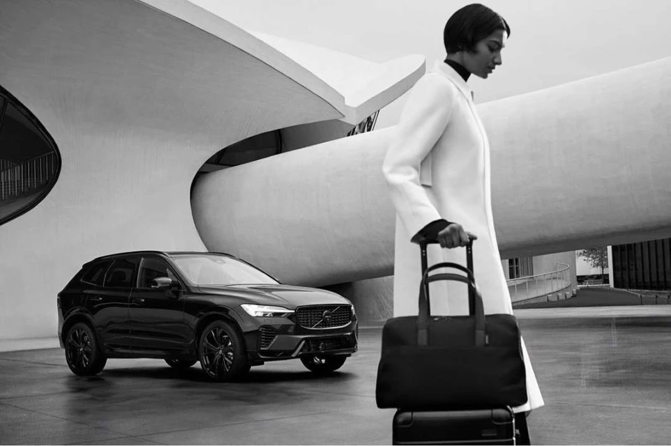 A modern black Volvo SUV and woman with a rolling bag stand before an architectural building in a monochrome outdoor shot.