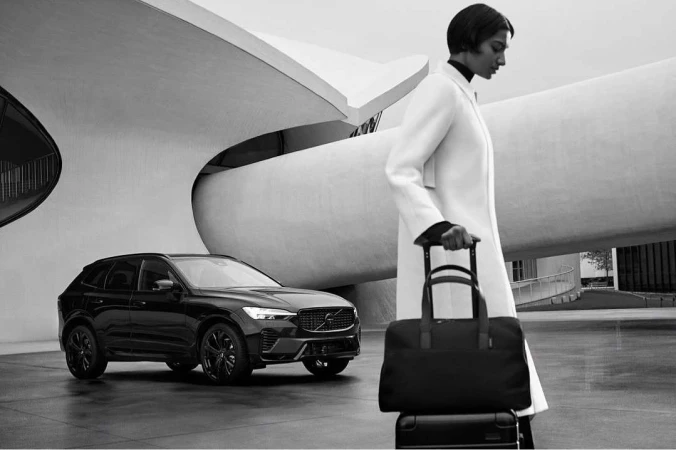 A modern black Volvo SUV and woman with a rolling bag stand before an architectural building in a monochrome outdoor shot.