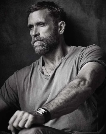 Monochrome portrait of a bearded man in his 40s wearing a t-shirt against a dark textured backdrop