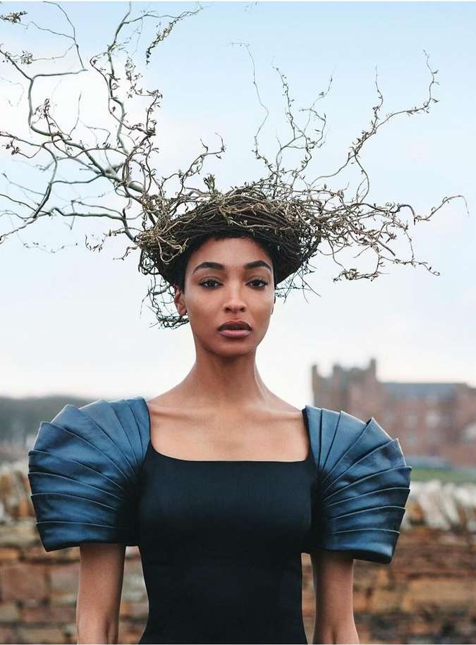 Artistic portrait of a woman with striking shoulder piece and a decorative headdress of twigs, set against a landscape backdrop.