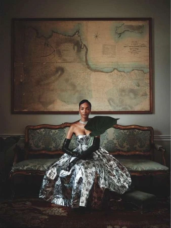 Woman in a strapless metallic patterned gown and black opera gloves seated on a vintage sofa under a framed map