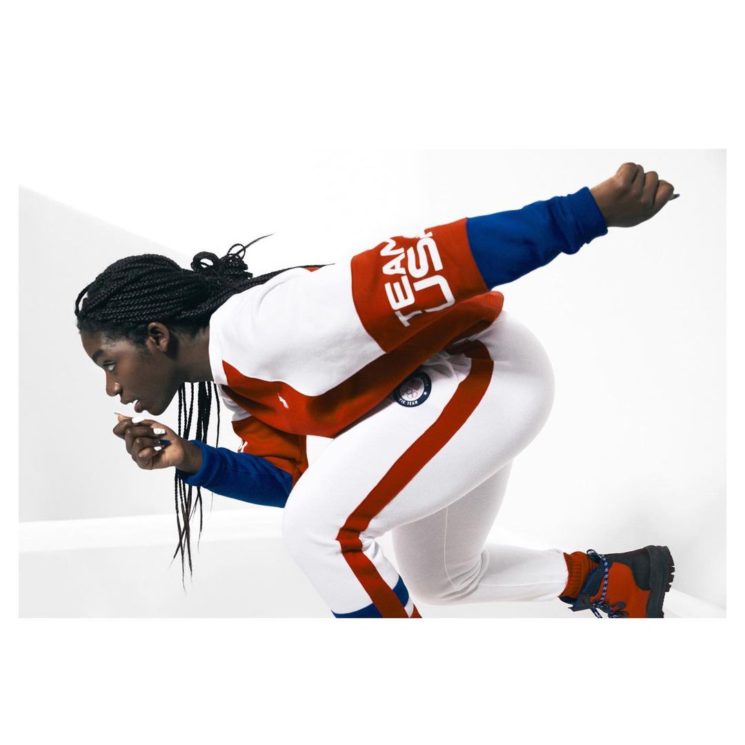 An African-American woman is captured in action, dressed in a white, red, and blue sweatsuit, ready to go, set against a clean white backdrop.