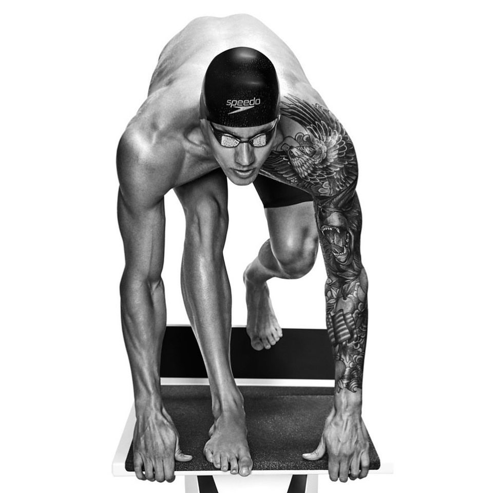 Monochrome shot of a muscular swimmer poised on the starting block, face visible behind goggles, showcasing detailed arm tattoos.
