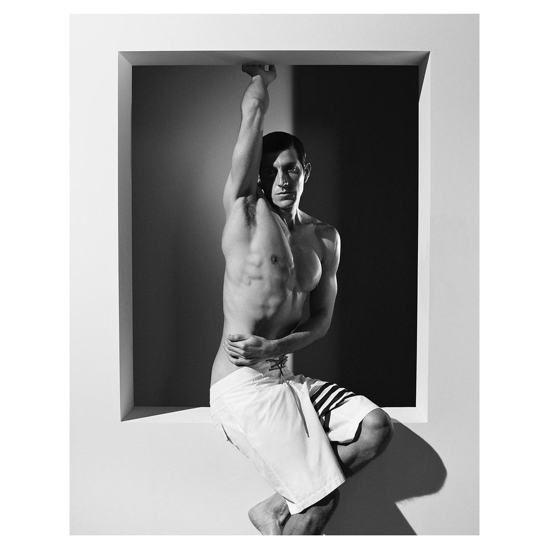 Athletic man in white swim trunks hangs in a white framed square, against a grey and black background. Studio lighting, monochrome.