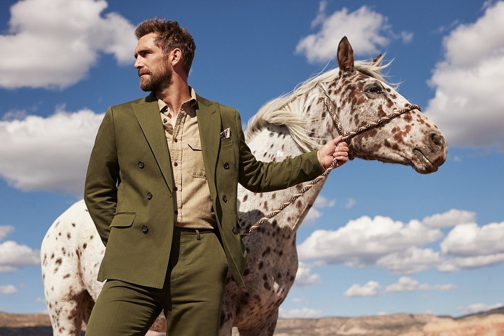 Adult man in olive green suit standing next to an Appaloosa horse in an outdoor desert setting with a cloudy sky backdrop.