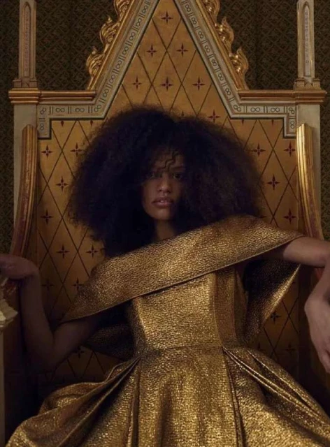Woman with afro poses in golden gown on a golden throne, against dark wallpaper for fashion editorial shot, eye-level angle.