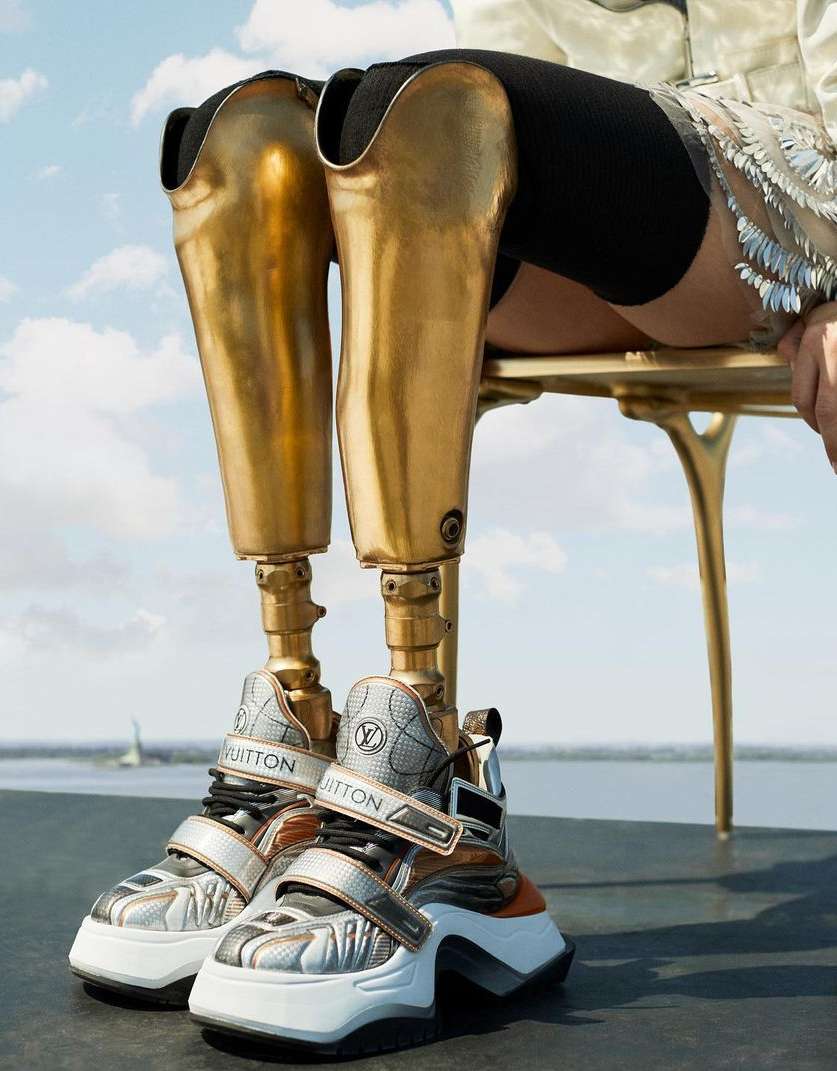 Fashion-forward studio shot of an individual with prosthetic legs wearing modern shoes, seated on a gold chair against a sky backdrop.