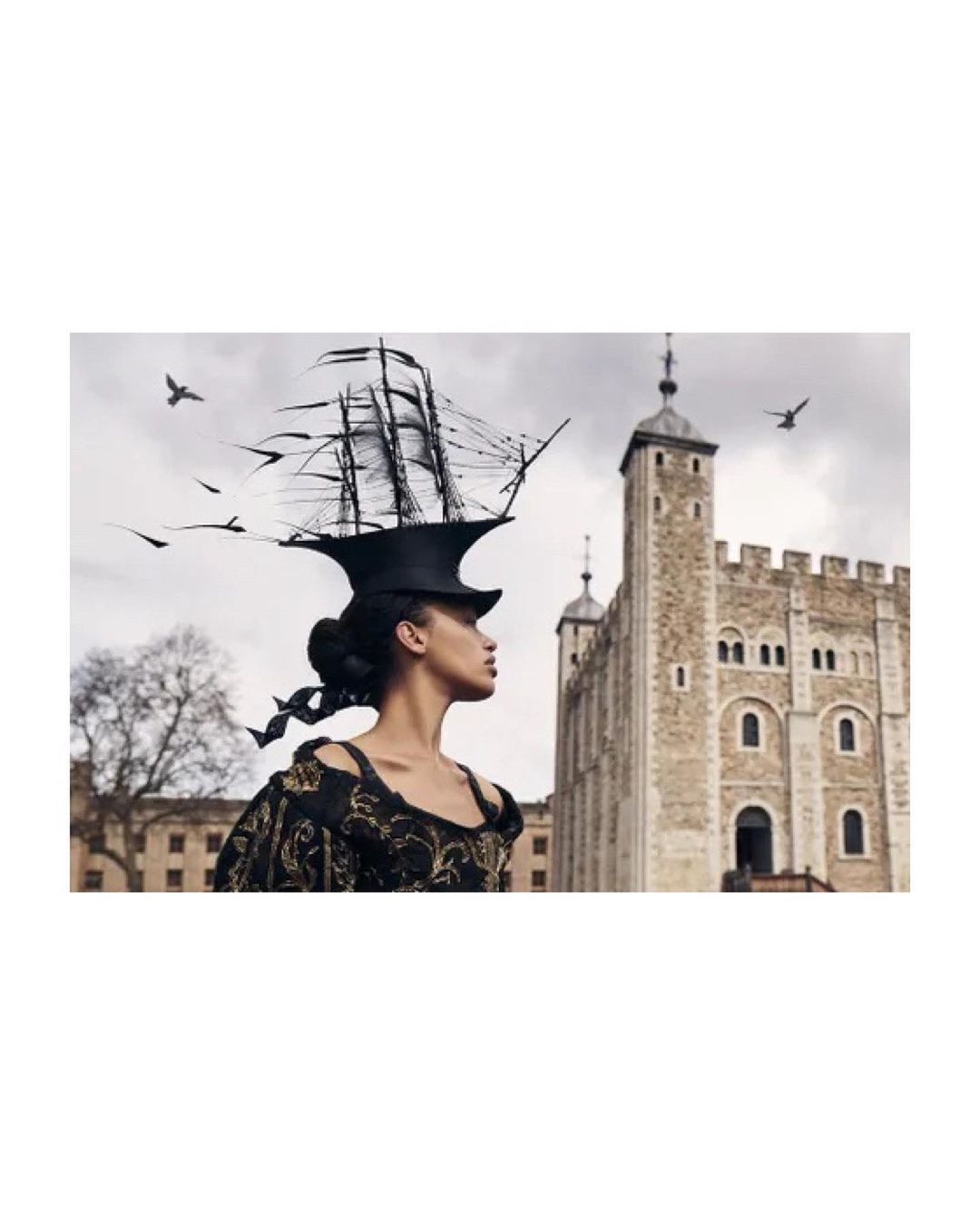 A woman in period clothing and a ship hat stands in front of a tall stone tower on a cloudy day with a few birds flying nearby.
