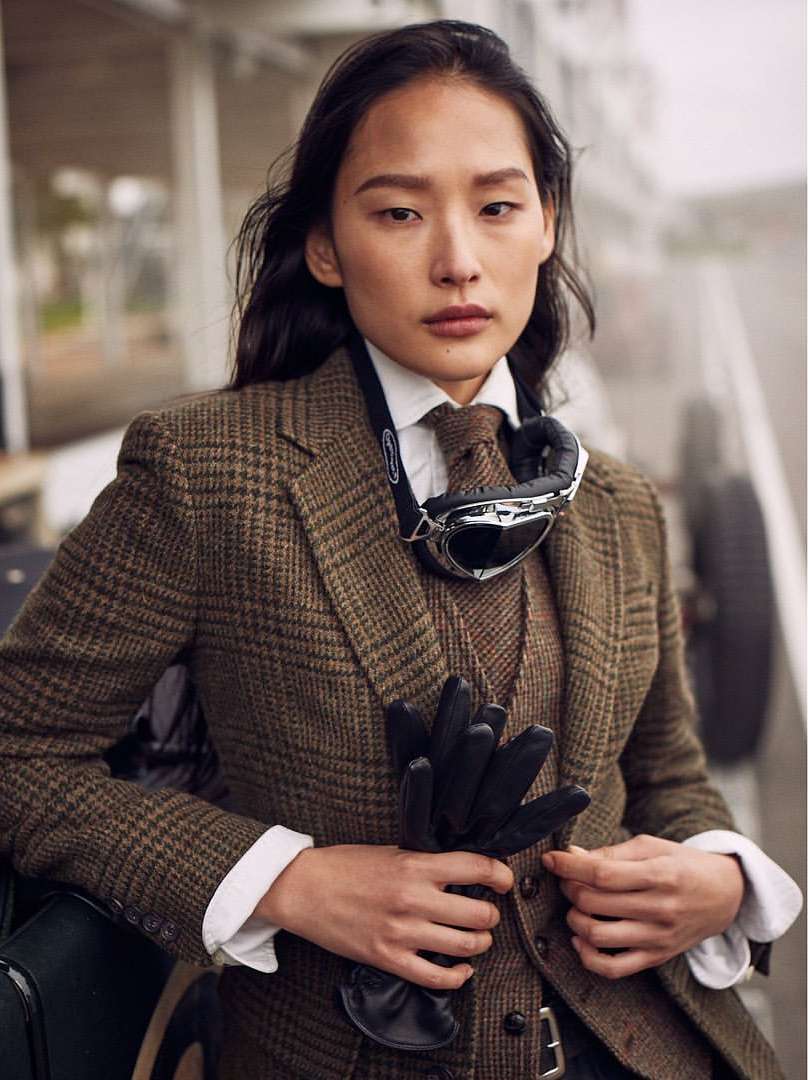 Elegant woman in tweed blazer, vest, and tie, wearing aviator goggles, holding gloves, stands outside near a classic car on an overcast day.