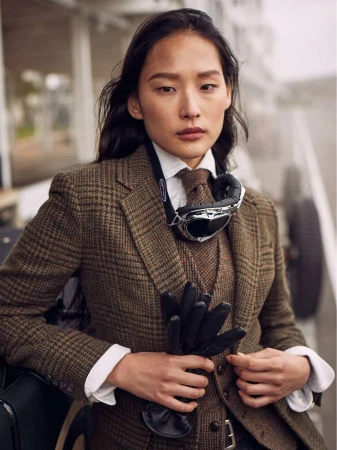 Elegant woman in tweed blazer, vest, and tie, wearing aviator goggles, holding gloves, stands outside near a classic car on an overcast day.
