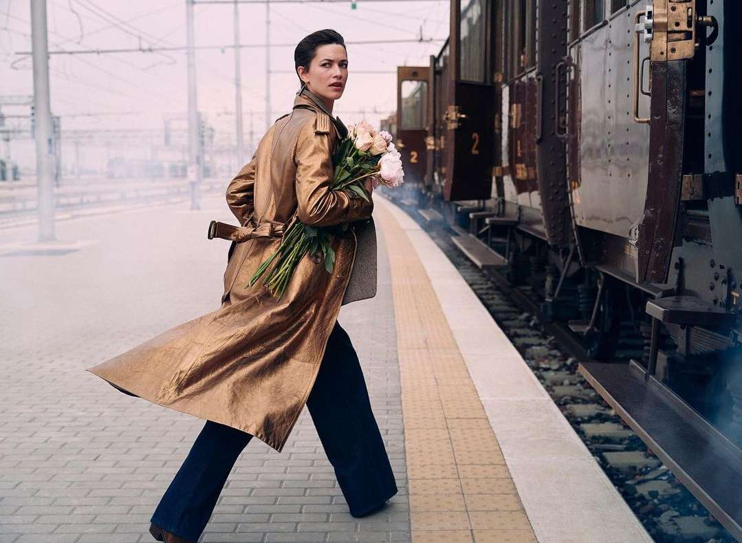 Woman in trench coat crosses train platform with flowers. Vintage train car, urban setting, and elegant editorial fashion.