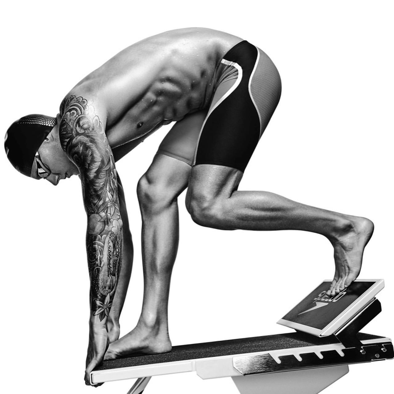 Black and white image of a muscular swimmer poised on the starting block wearing a cap and goggles, tattoos on his arm, ready to dive.