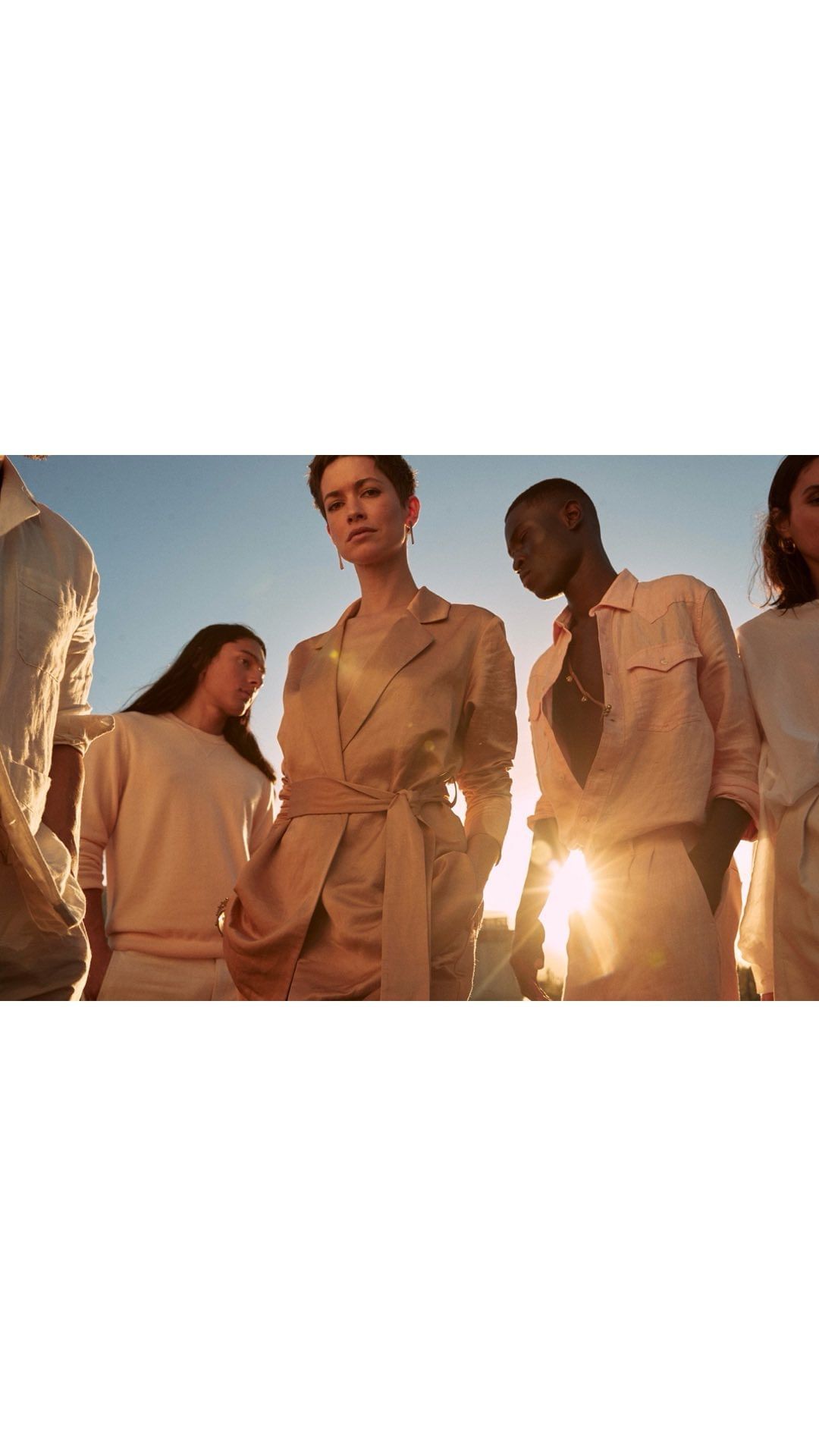 Diverse group of young adult models in sunlit outdoor fashion shoot wearing neutral linen clothing, captured at a low angle.