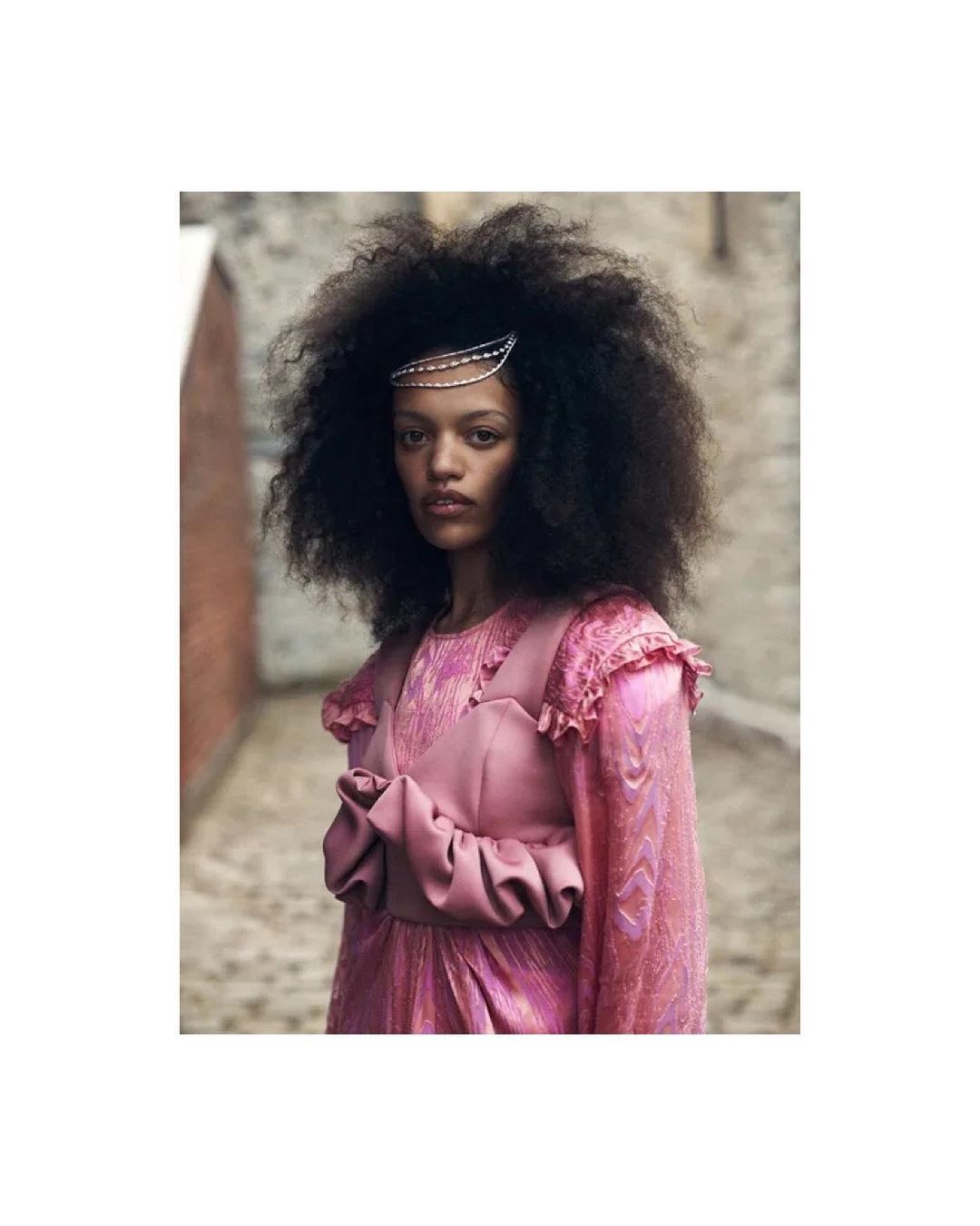 A woman with natural hair in a pink dress poses outdoors. This eye-level shot highlights her vintage-inspired fashion and hair accessory.