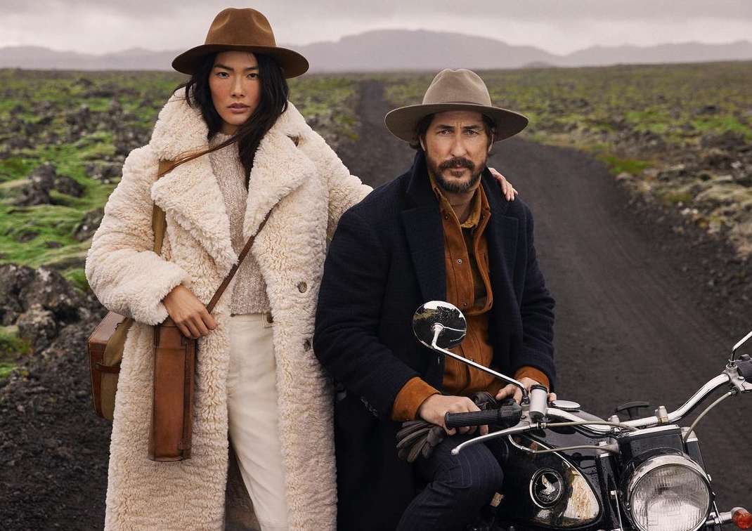 Man and woman outdoors on a dirt road, the woman standing in a fluffy white coat and the man seated on a motorcycle.
