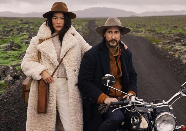 Man and woman outdoors on a dirt road, the woman standing in a fluffy white coat and the man seated on a motorcycle.
